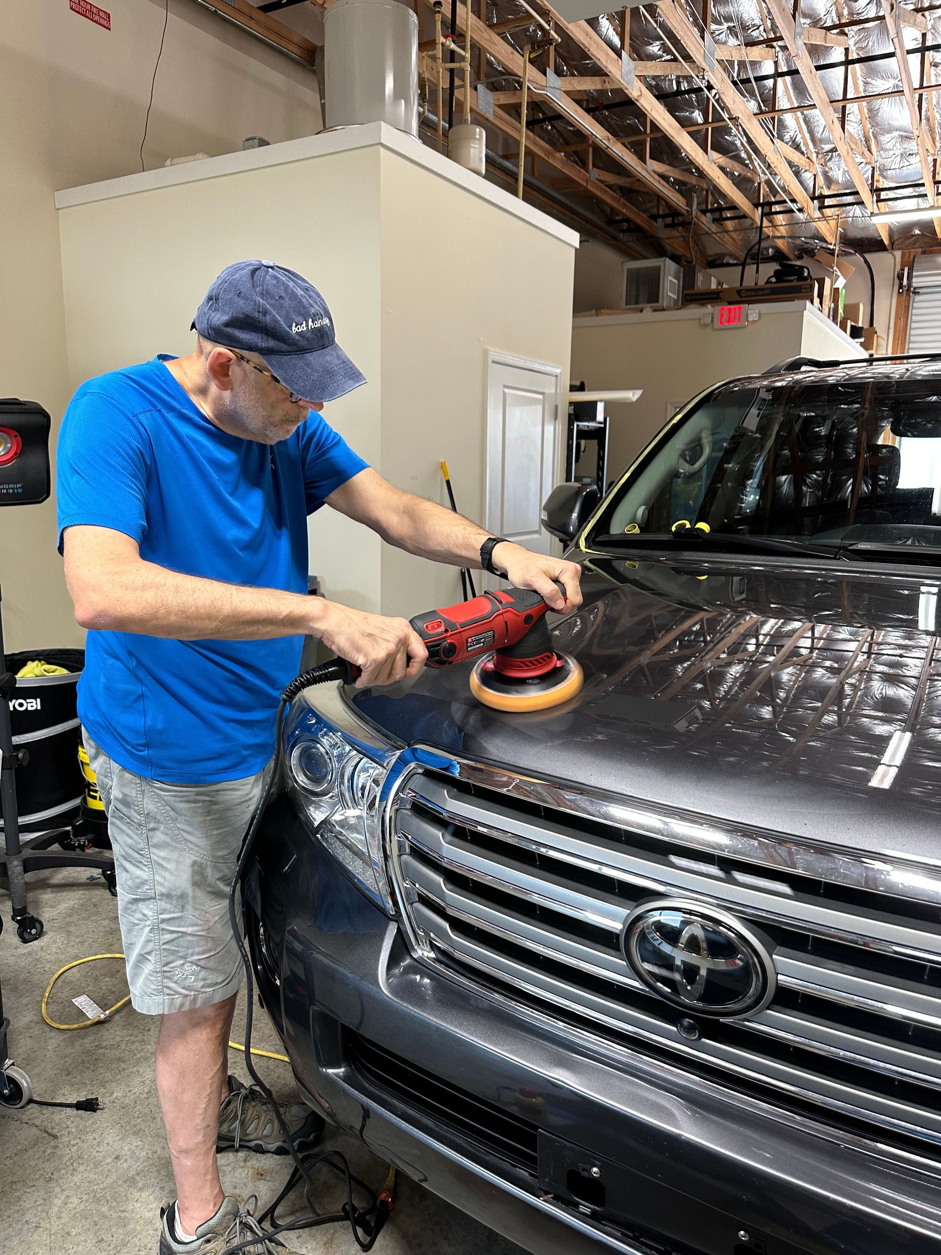 Man polishing the hood of a dark gray SUV with a red and black power buffer in a garage.