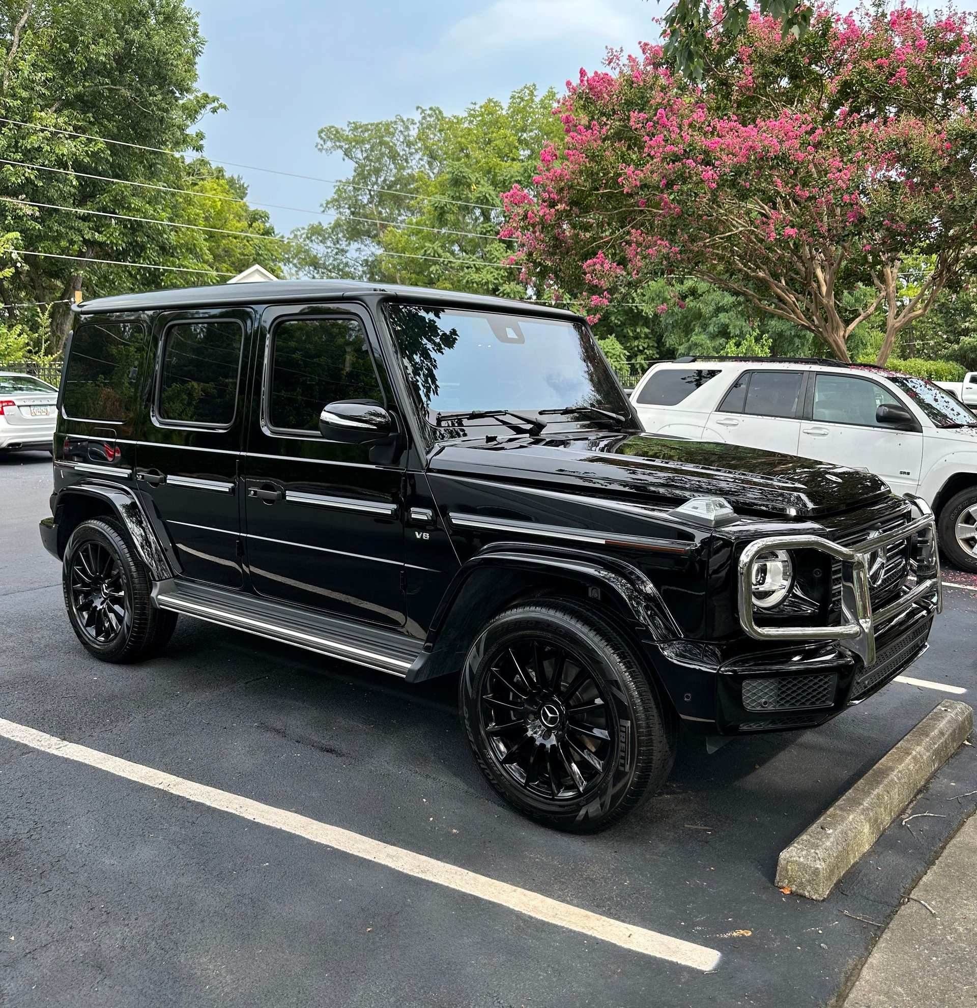 Black Mercedes-Benz G-Class SUV parked in a lot, with tinted windows, black wheels, and a tree in the background.