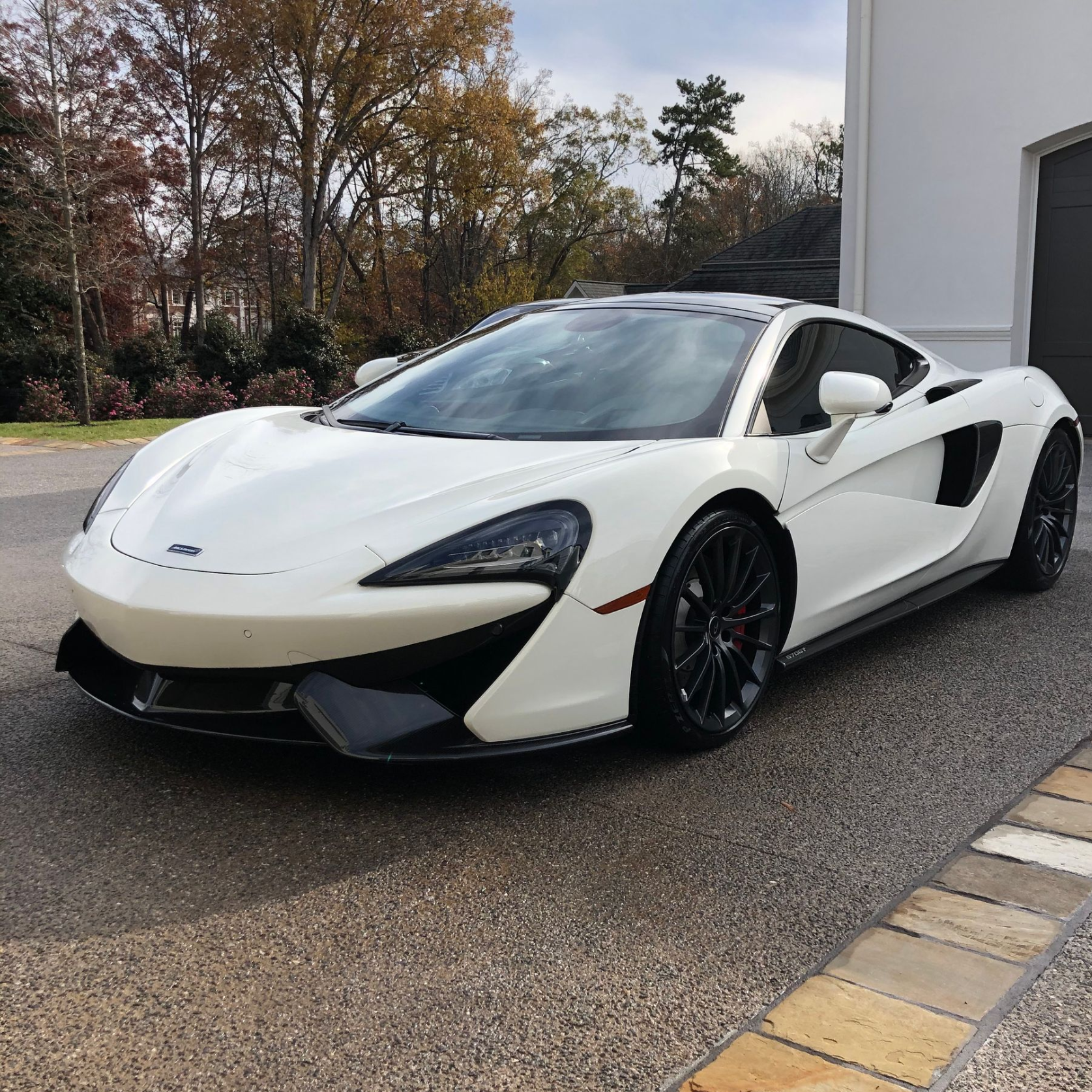 White McLaren sports car parked on a paved driveway in front of a building with a closed garage door.