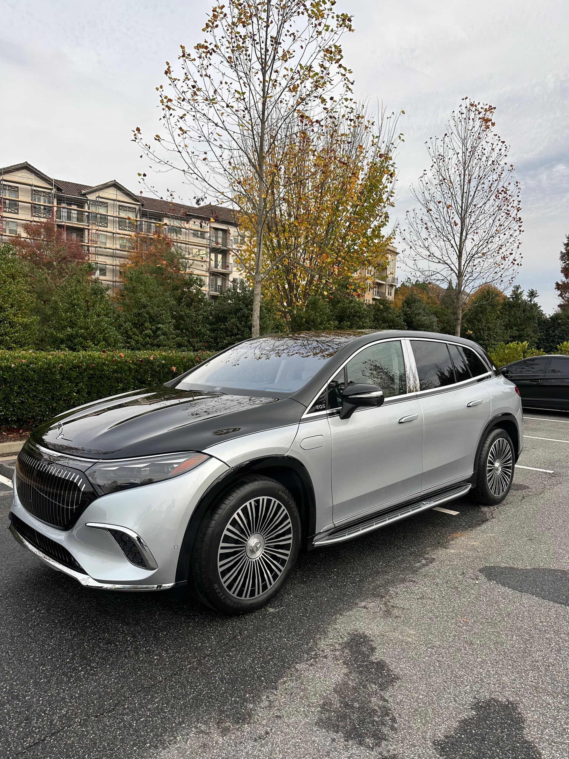 Silver and black Mercedes EQS SUV parked in a parking lot, with a building and trees in the background.