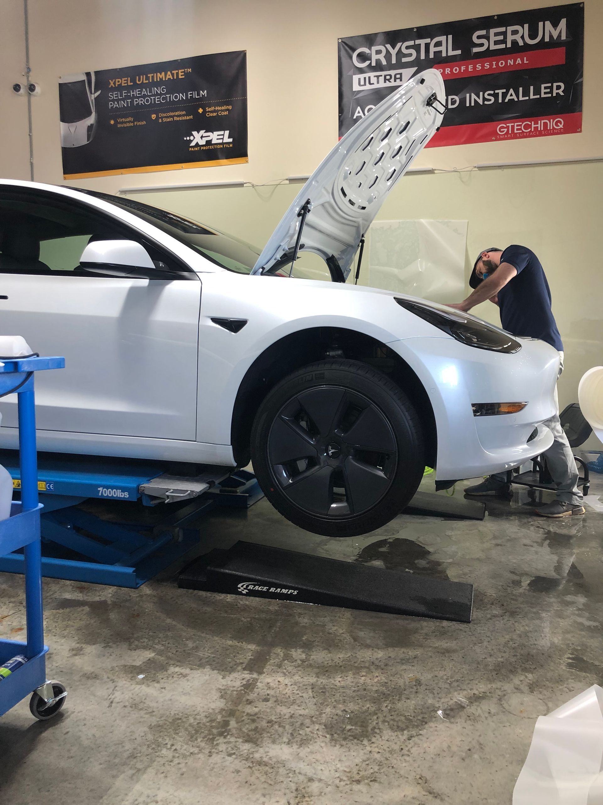 A mechanic working on a white Tesla with its hood open in a garage. Car is on a lift.