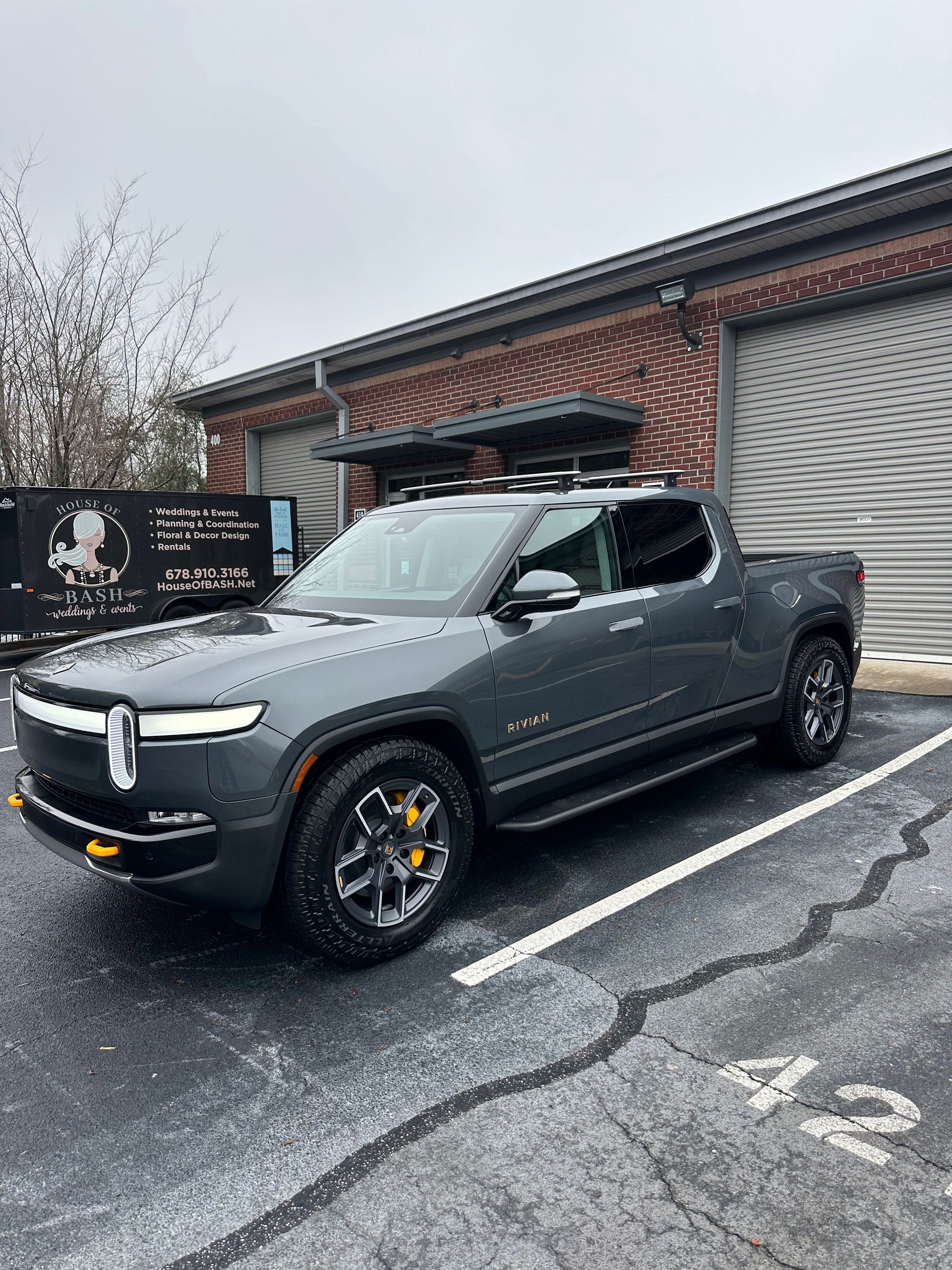 Gray Rivian pickup truck parked near a brick building. It has a roof rack and yellow brake calipers.