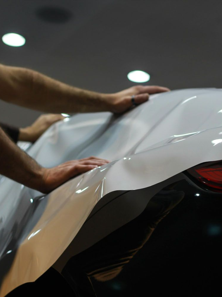Hands smoothing a clear wrap over the rear of a white car, likely for protection.