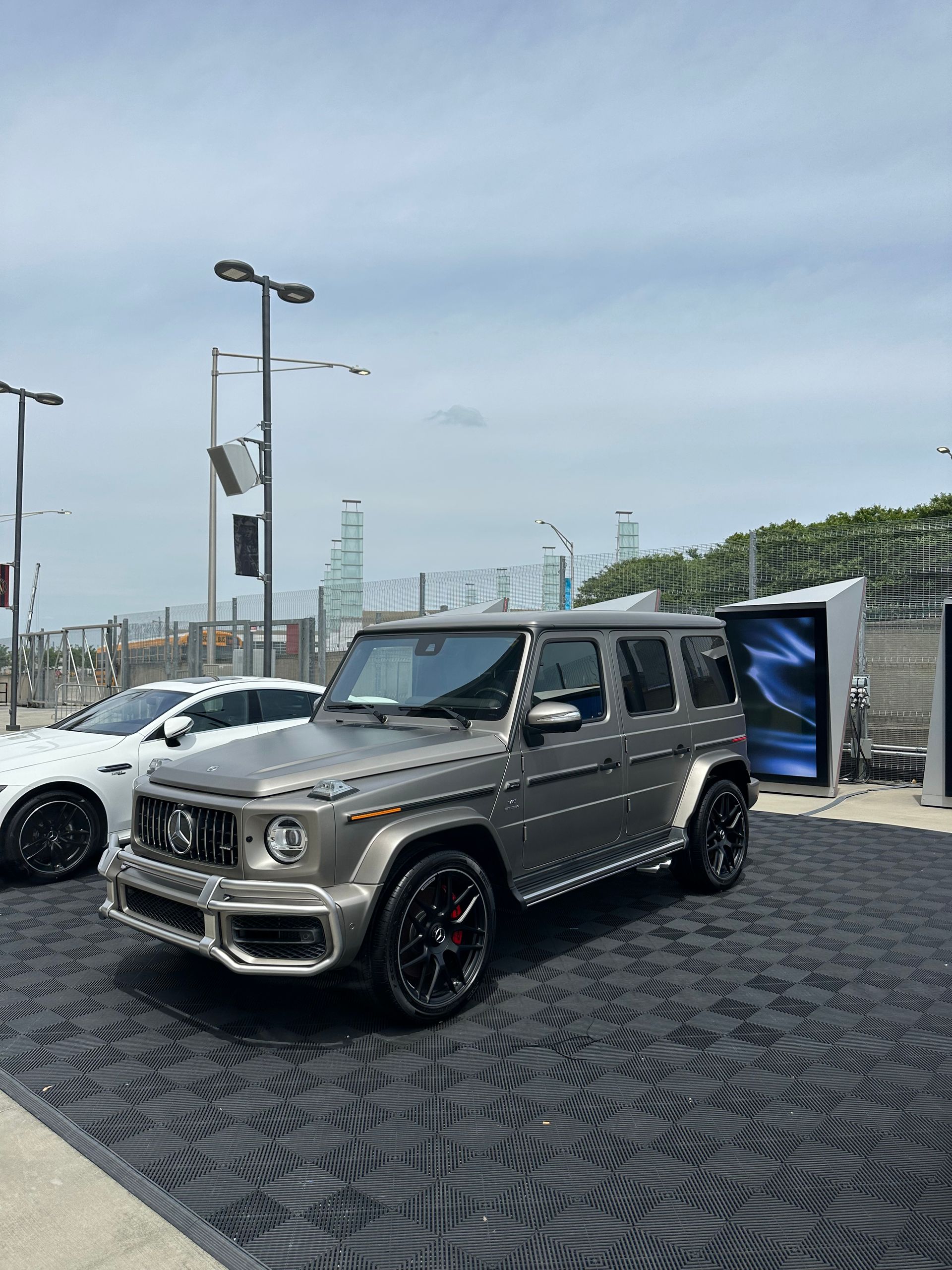 Gray Mercedes-Benz G-Wagon SUV on black mat; white car visible. Outdoors, under a cloudy sky.