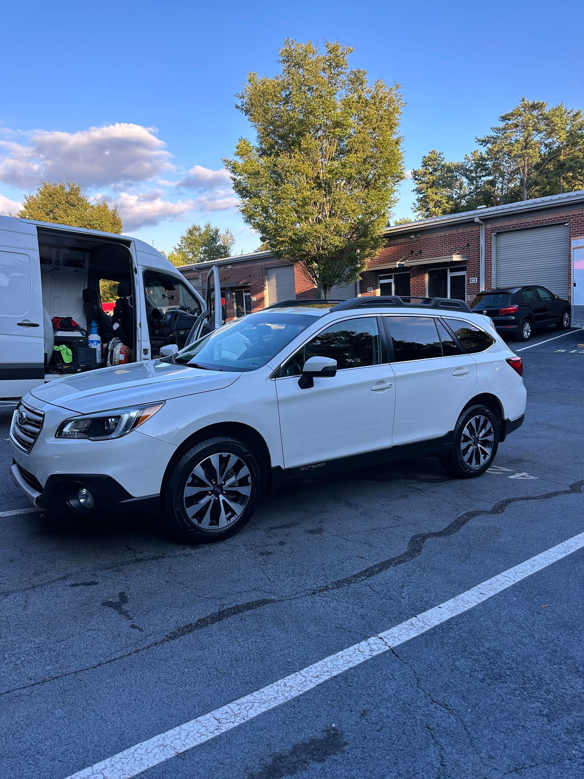 White Subaru Outback parked on asphalt, near a white van and brick building on a sunny day.