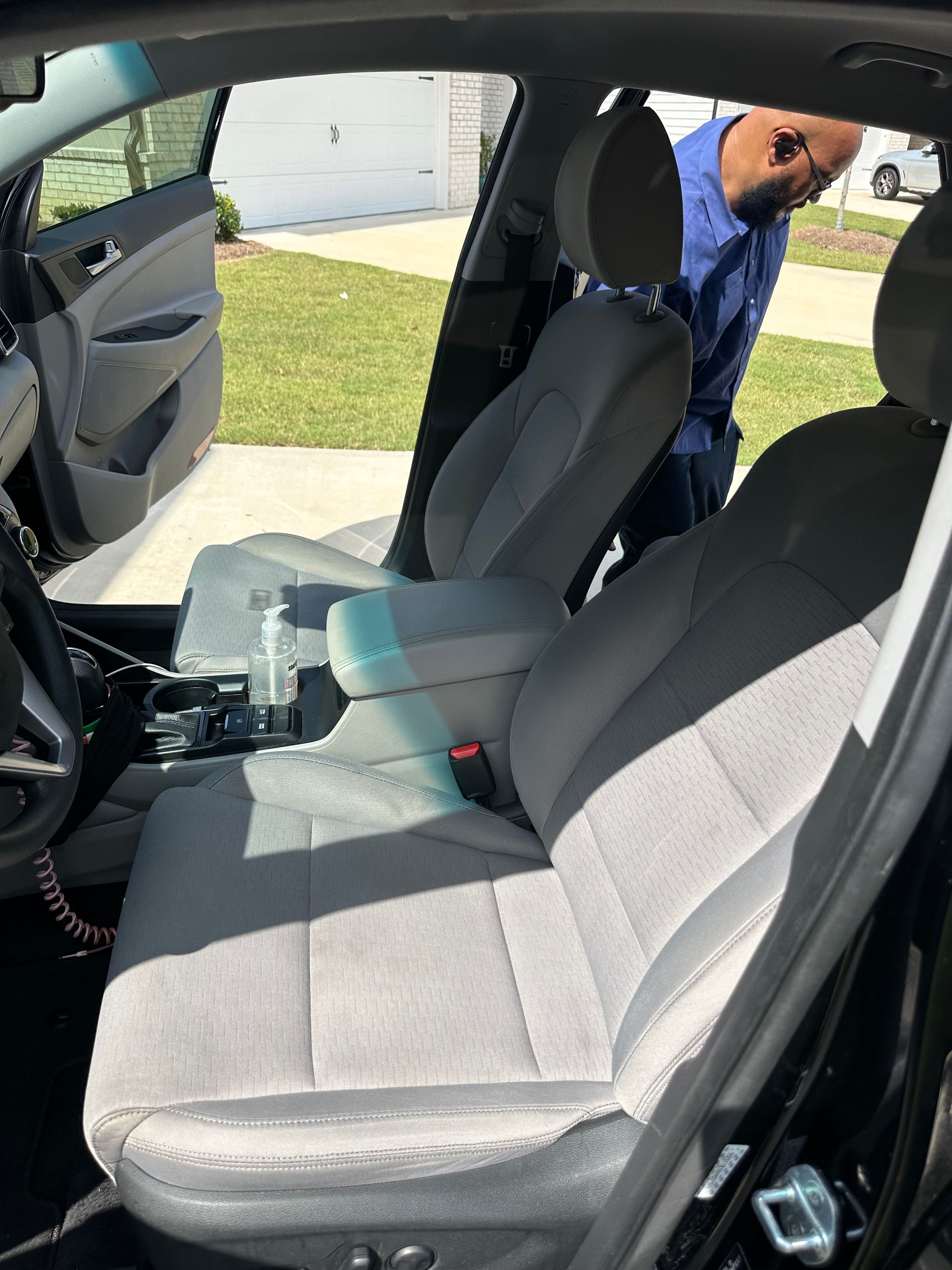 Interior of a black car with gray seats. A person stands near the open driver's side door, looking inside. Sunlight.