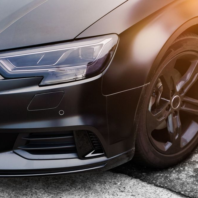 Black car's front with a headlight, a wheel, and a black rim. Sunlight on the right side.
