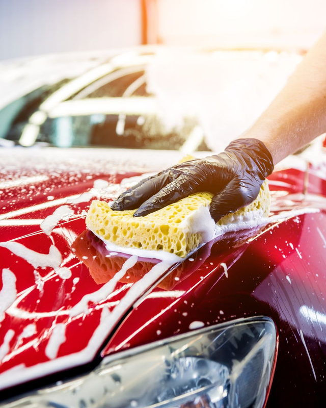 Gray car being washed with a hose, foam covering the body and wheel on a wet driveway.