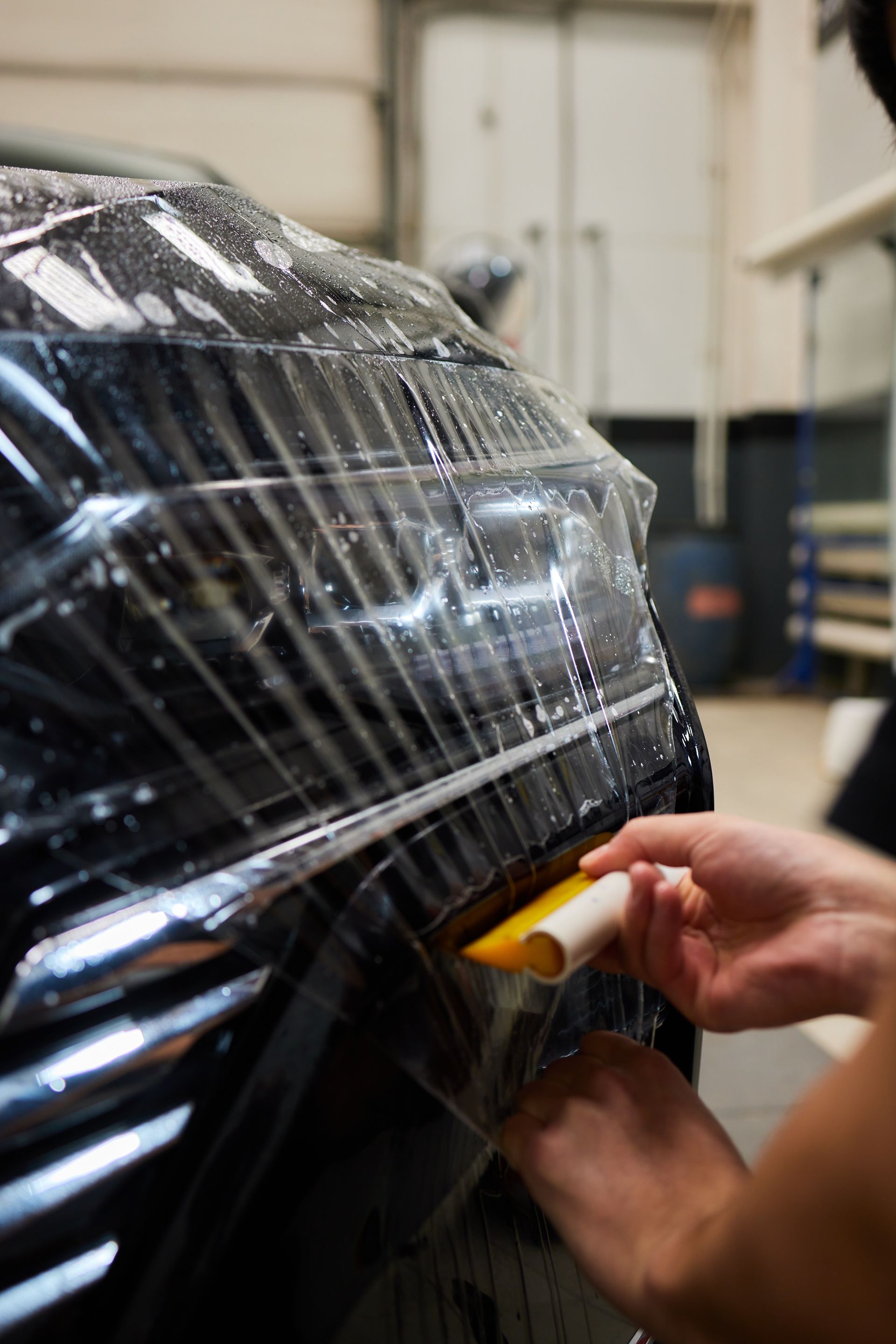 Person applying clear protective film to a car's headlight with a yellow squeegee.