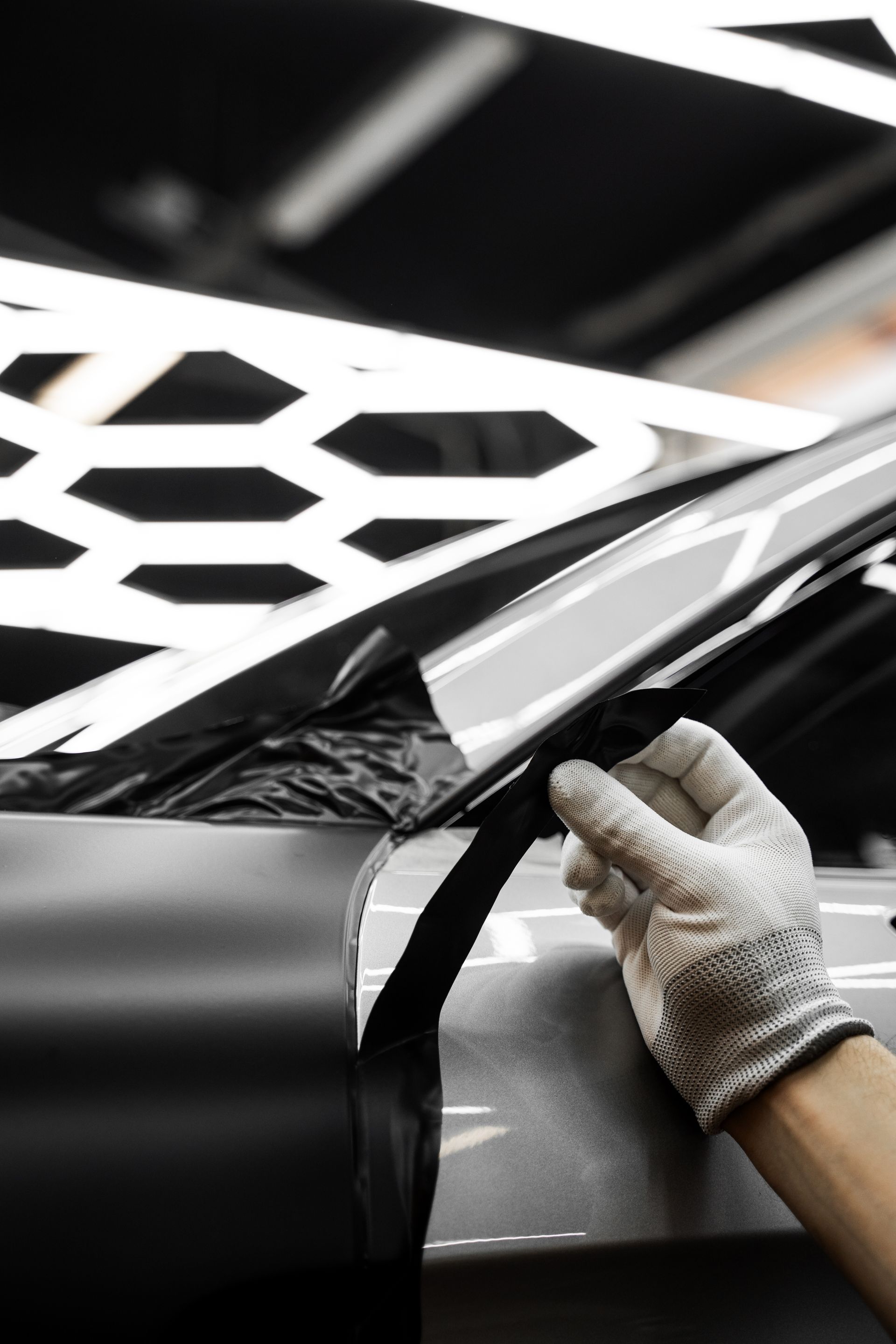 Gloved hand peeling back black vinyl on a car. The vehicle is silver. Bright lights shine above.