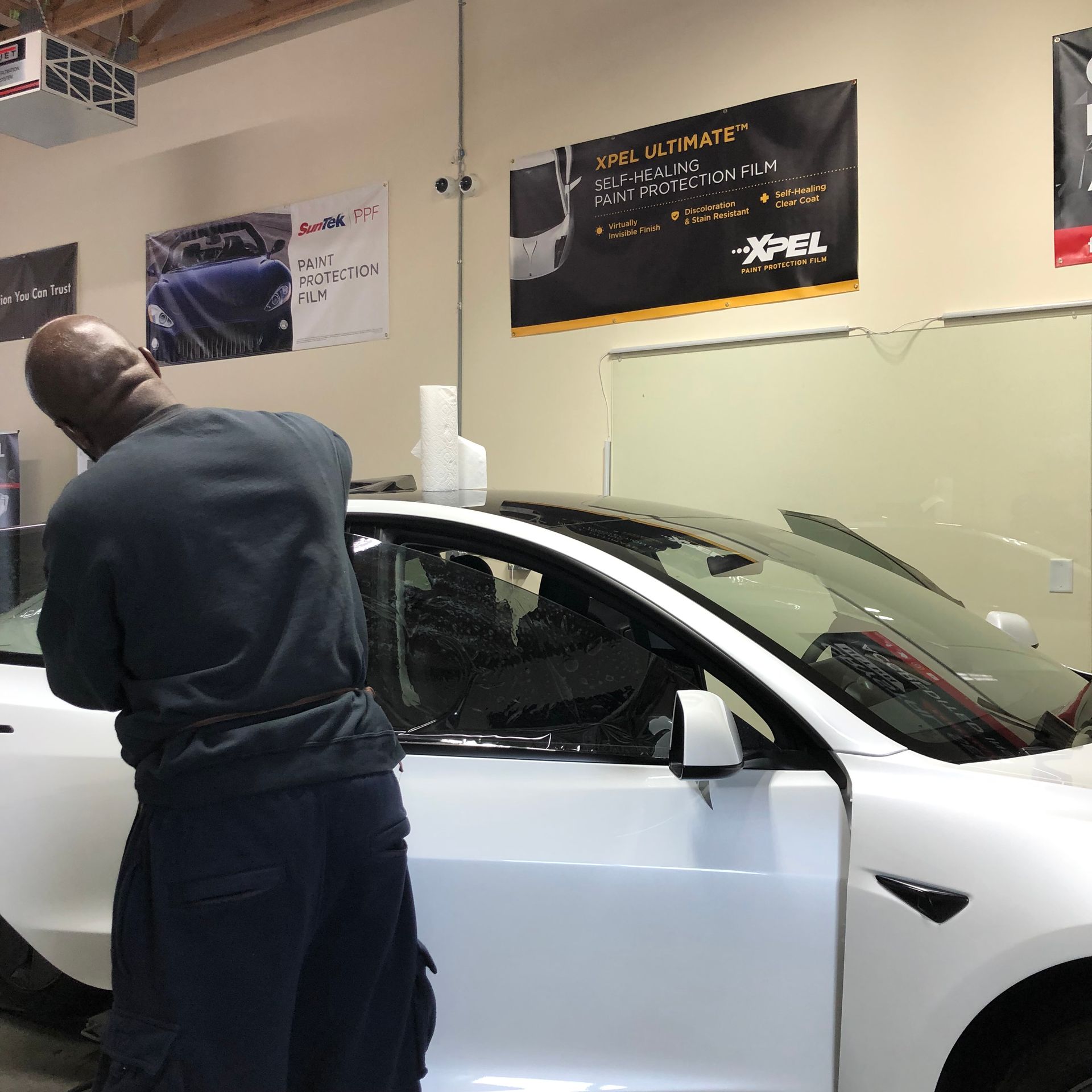 Man applying tint to a white Tesla in a garage, with banners and equipment in the background.