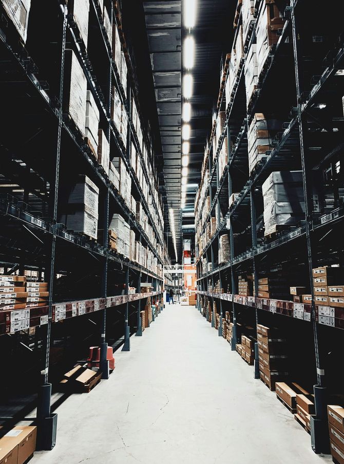 Warehouse aisle with shelves packed with boxes and goods, overhead lighting.