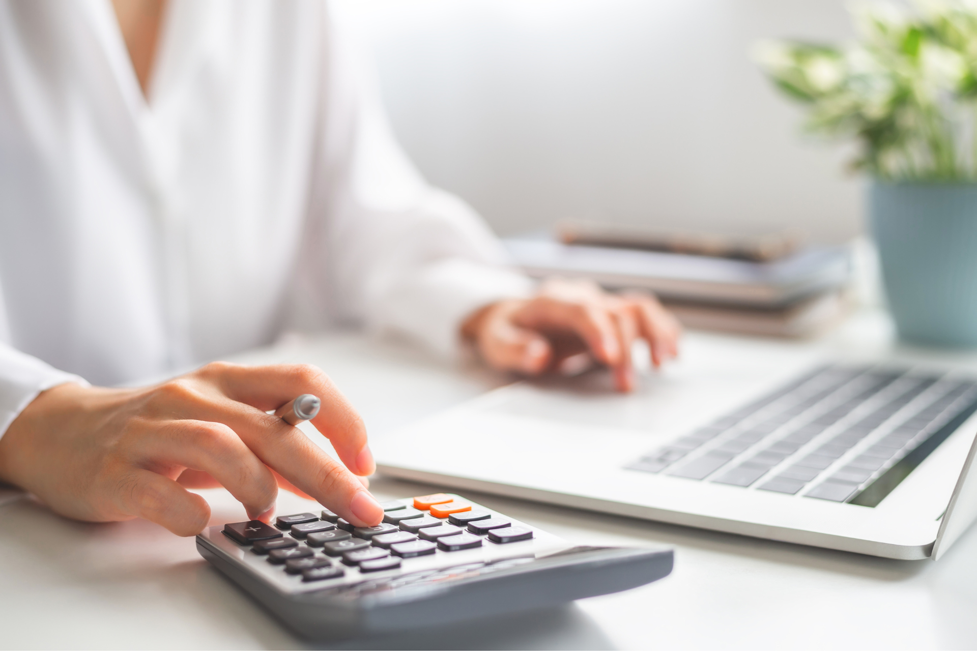 Les mains d'une femme utilisant une calculatrice sur un bureau blanc avec un ordinateur portable et une plante en arrière-plan.