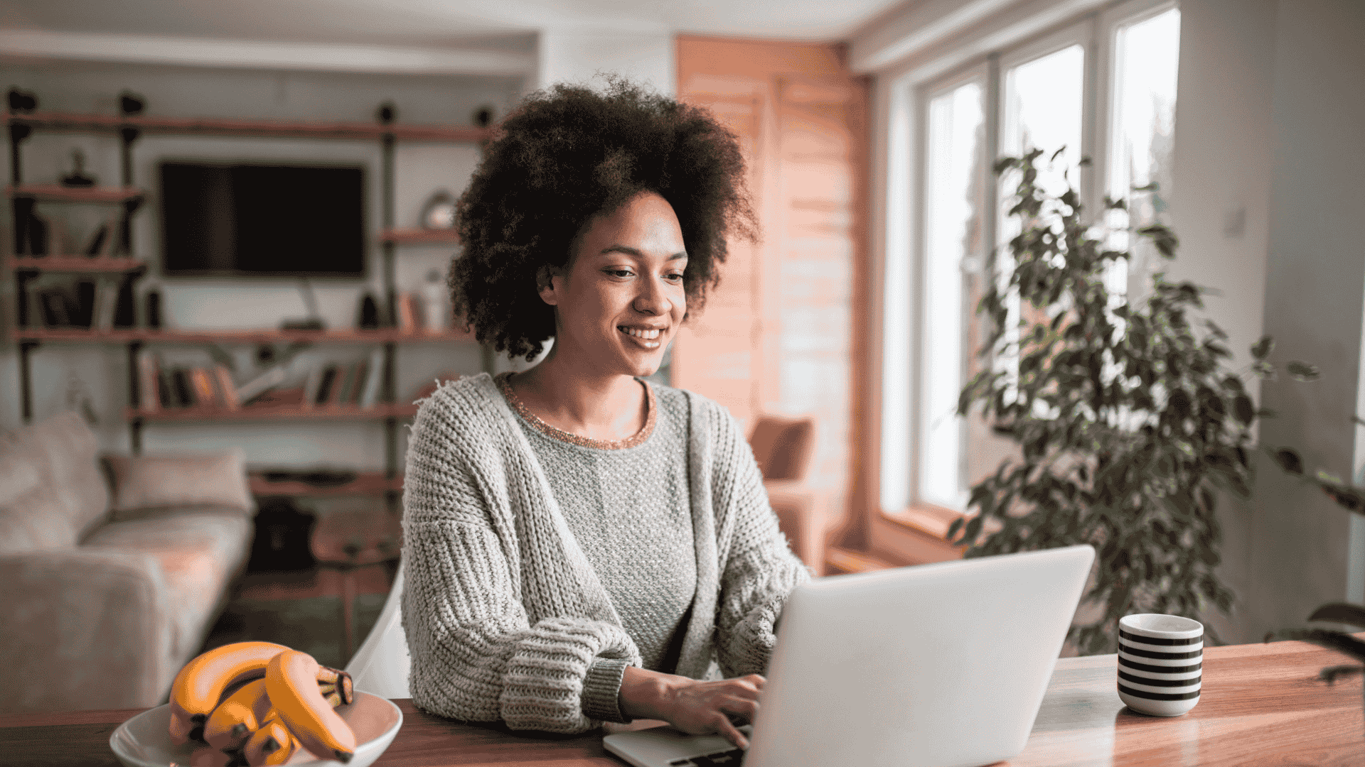 Femme afro souriante, travaillant sur son ordinateur portable à la maison. Bananes et café à proximité.