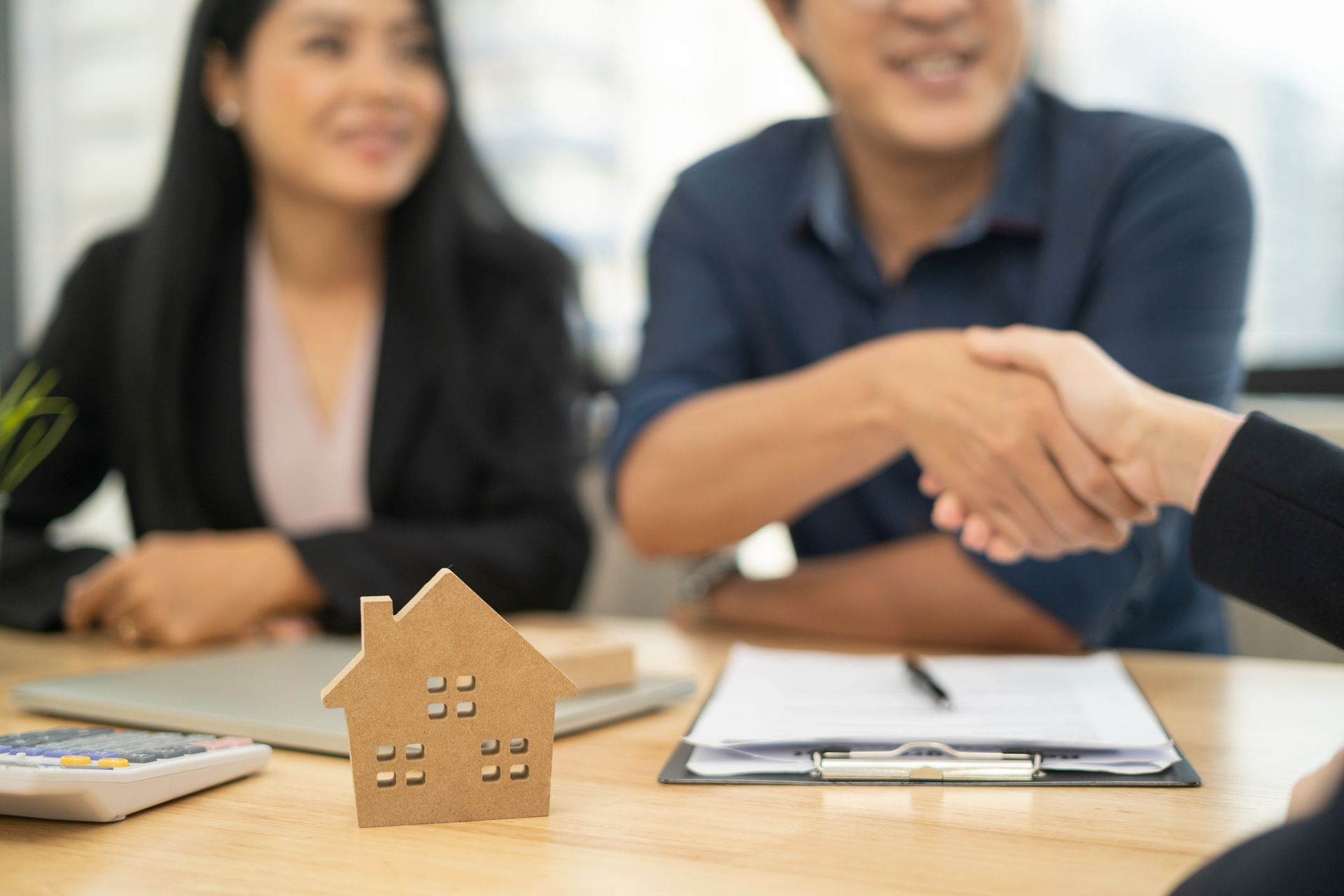 Couple shaking hands with a person at a table, wooden house model, paperwork.