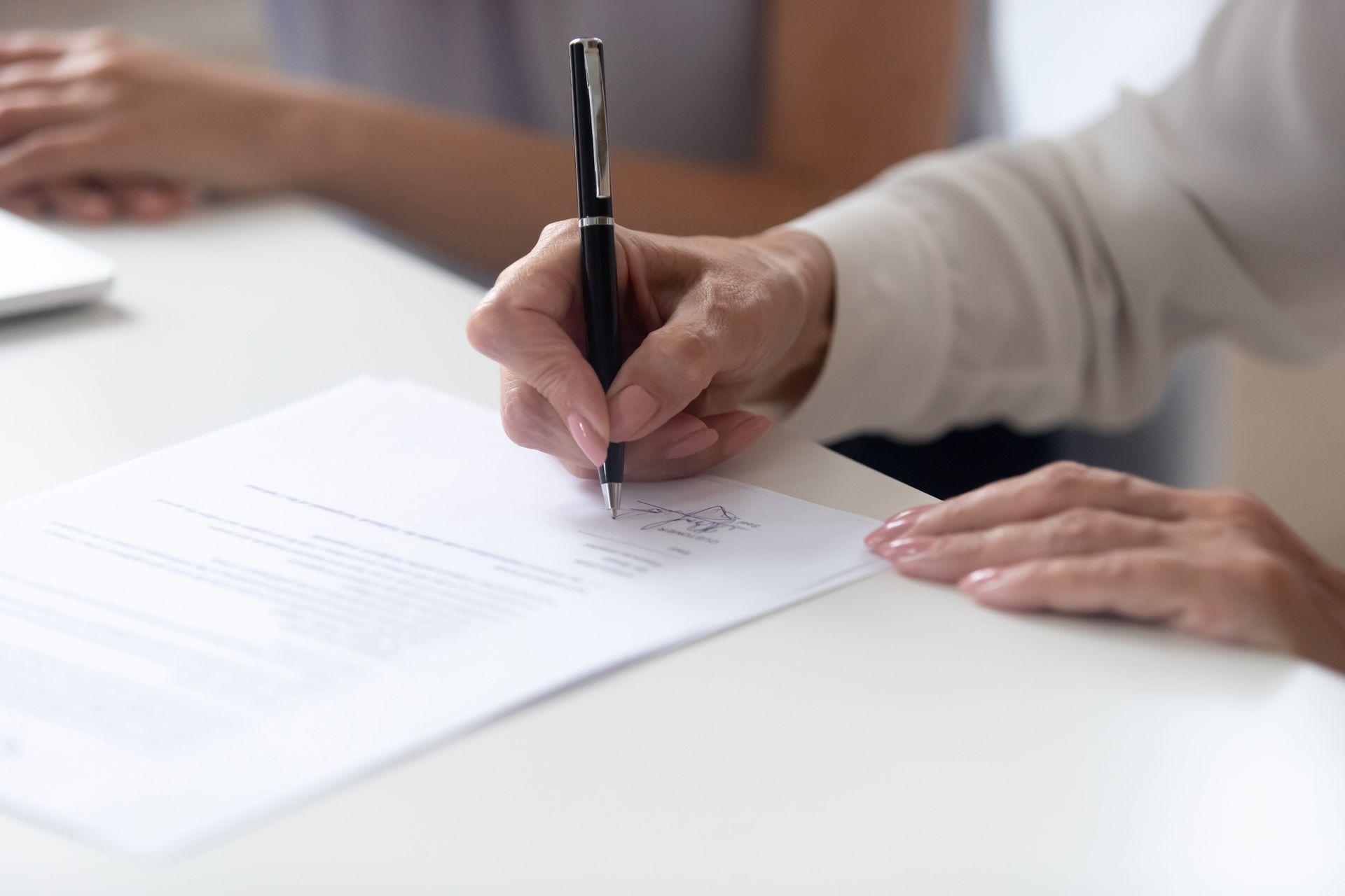 Person signing a document with a black pen on a white table. Another person's arm is visible on the left.