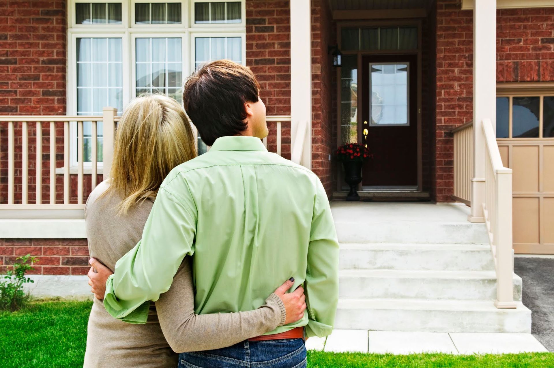 Couple gazing at a brick house, arms around each other. White porch, front door, garage.