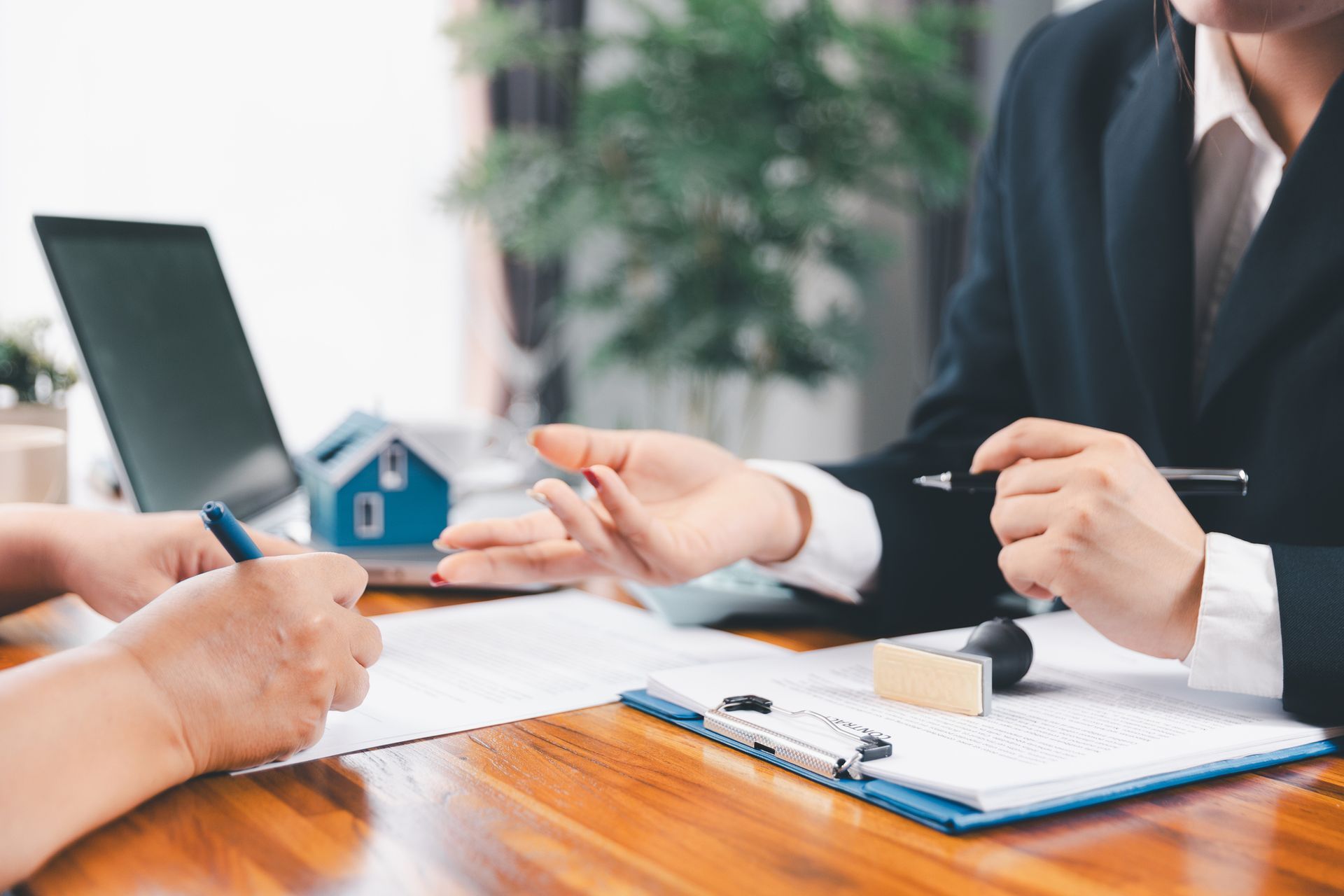 Two people at a desk review documents. One person signs, other gestures, next to a small house model and laptop.