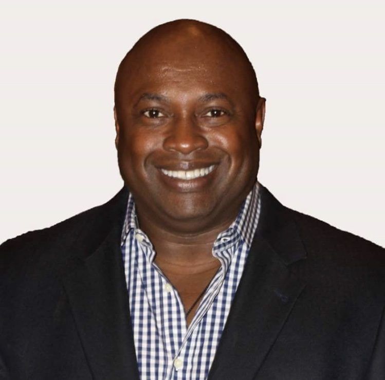 Man wearing a black blazer over a blue and white checkered shirt, smiling.