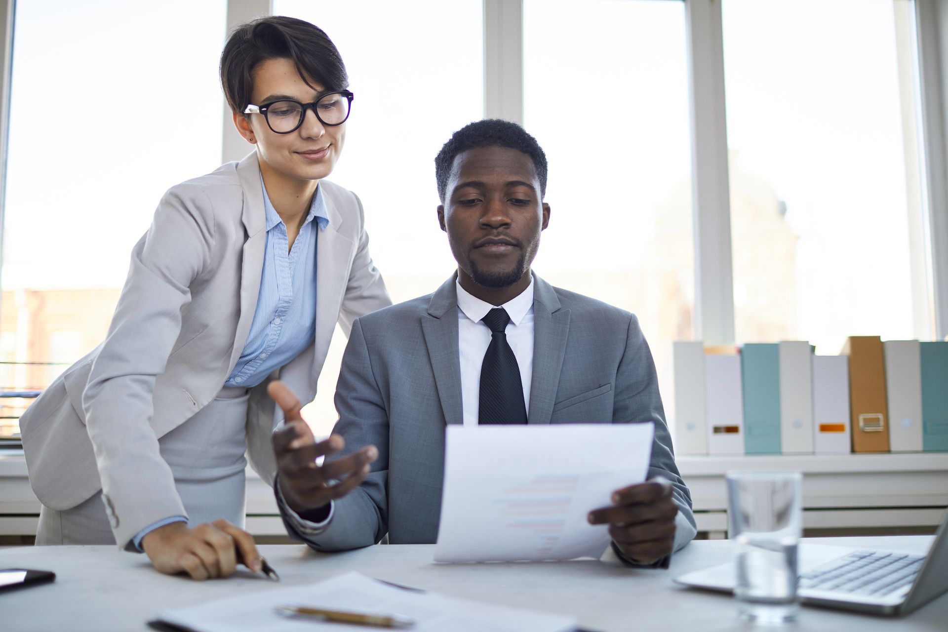 Woman in suit gestures at document with man in suit at desk.