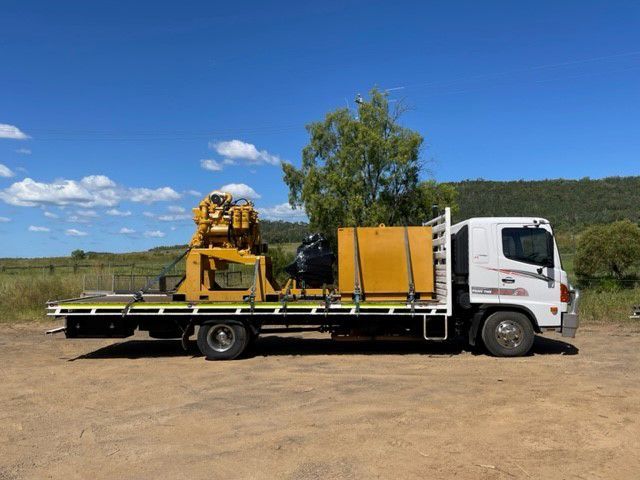 Yellow machinery on a flatbed truck, parked on dirt road with green hills and blue sky in the background.
