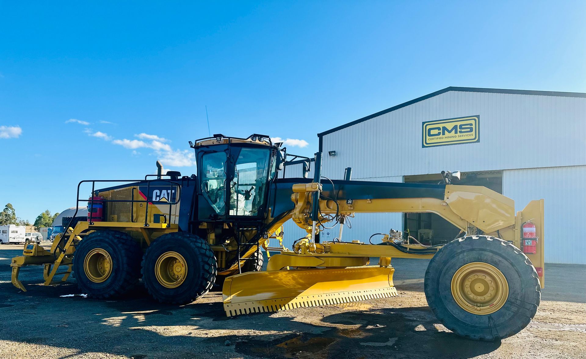 Yellow road grader on gravel with blue sky and clouds.