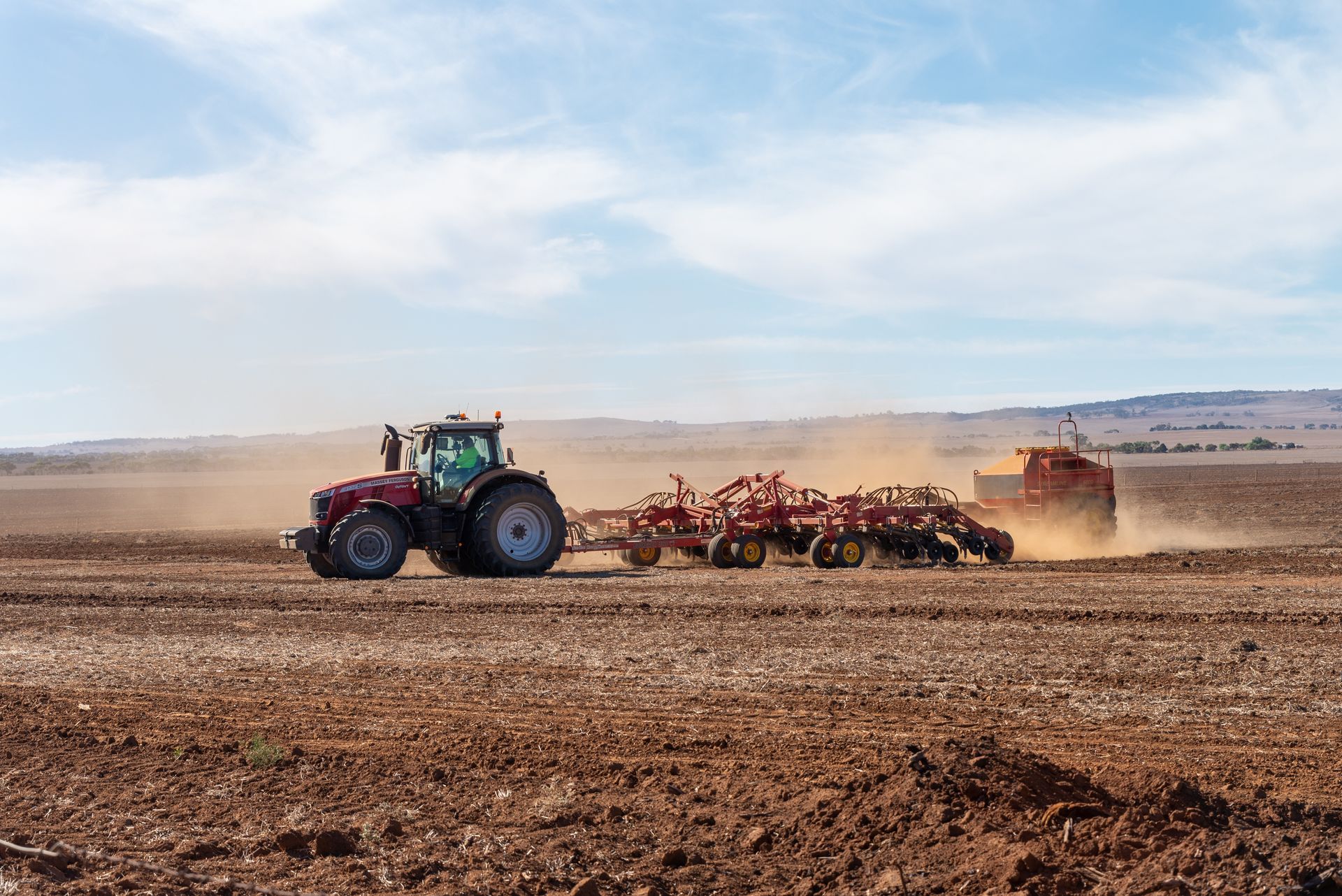 Tractor plowing a field; red machine kicking up dust under a blue sky.
