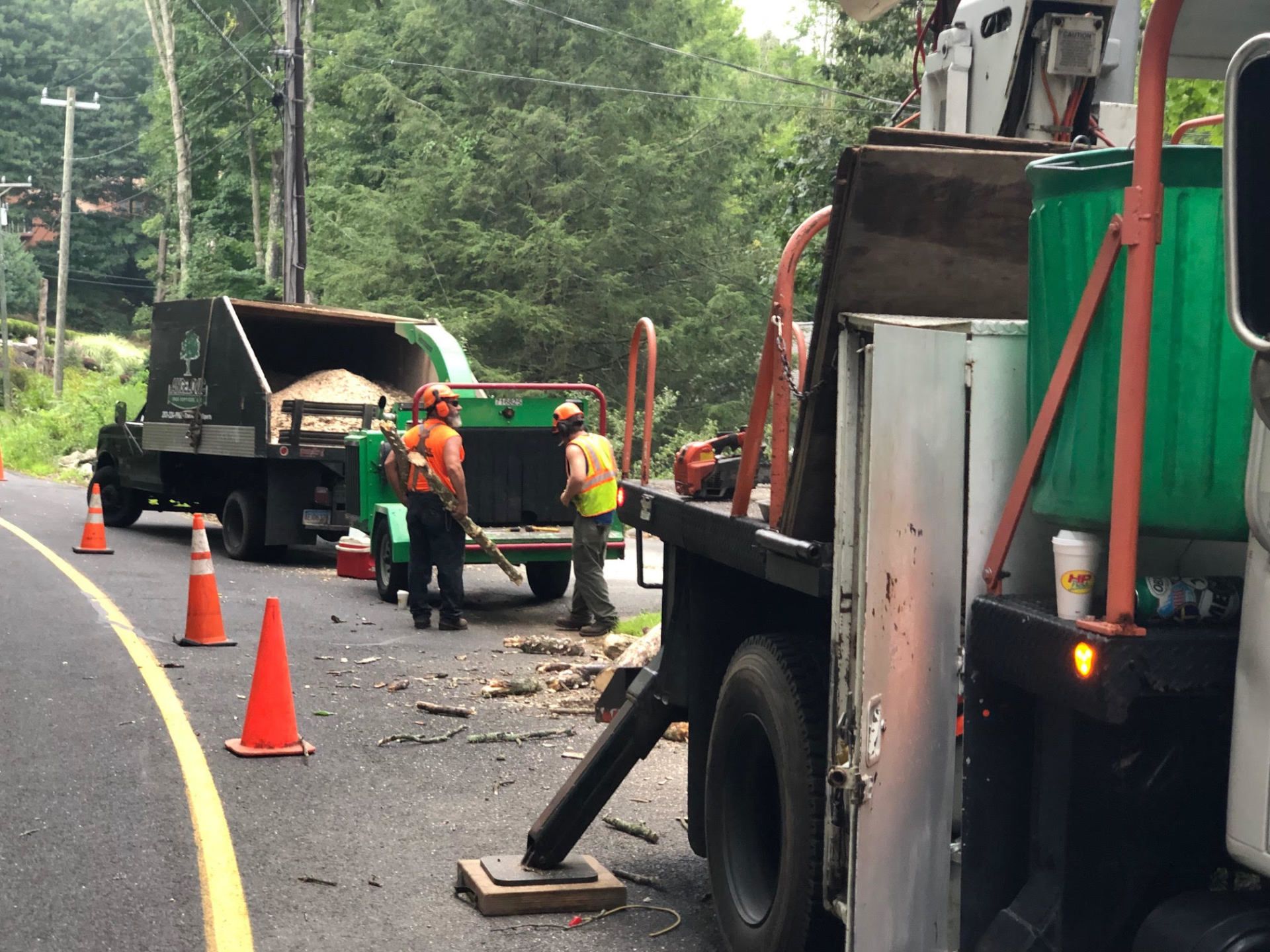 Workers chipping tree debris from a road with trucks and cones, likely for maintenance, outdoors.