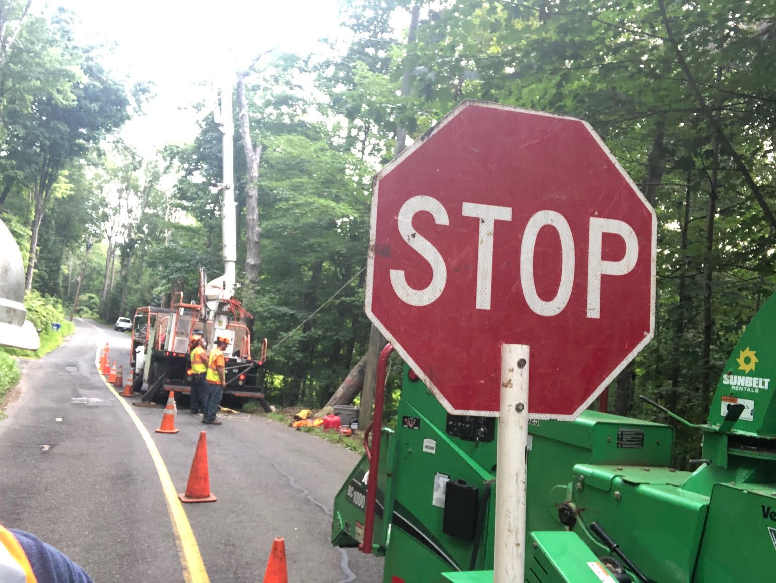 Stop sign near road closed for tree removal. Workers operate machinery.