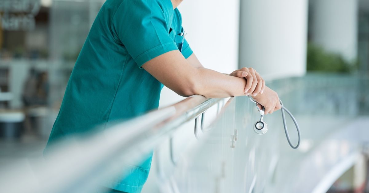 A medical professional is wearing scrubs and holding a stethoscope. They are leaning against a stainless steel handrail.