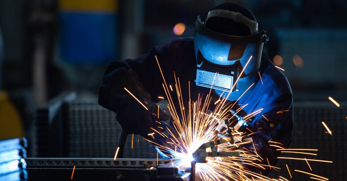 An industrial worker wearing protective clothing and a welding helmet while welding steel. Welding sparks fly through the air.