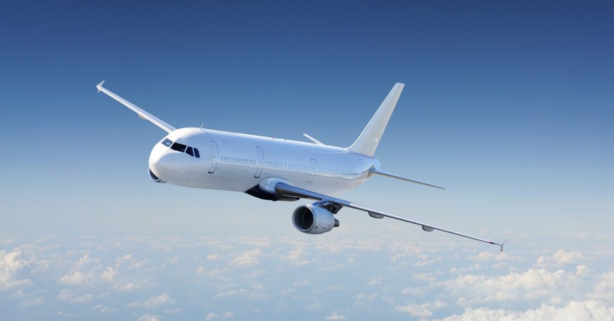 A white airplane flying through the blue sky on a sunny day. The plane flies well above pockets of clouds.