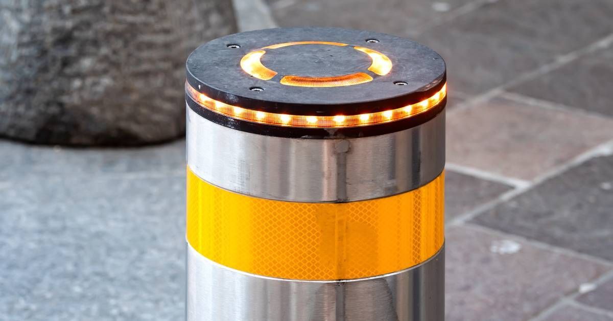 A close-up view of a stainless steel bollard with yellow reflective detail. The top of the bollard has a row of lights.