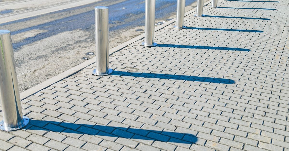 A row of stainless steel bollards lining an empty cobblestone sidewalk. Each bollard casts a long sh