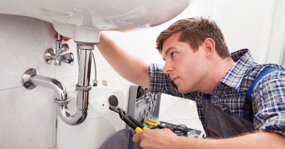 A man in a collared shirt and overalls working on the metal pipes beneath a white porcelain bathroom sink.