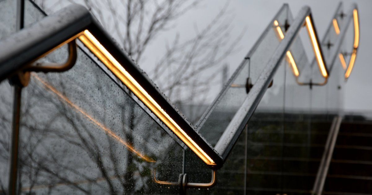 A close-up of an exterior stainless steel handrail with raindrops and glowing recessed LED strips on the underside.