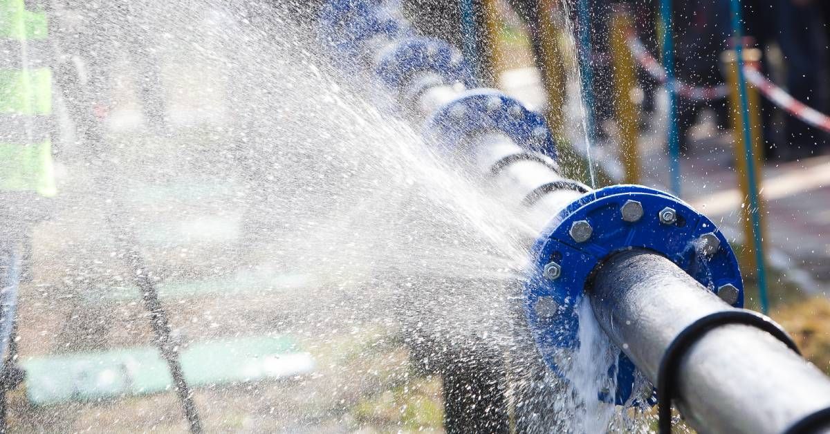 A burst steel pipe with several blue rings with bolts in them. The pipe is spraying water everywhere