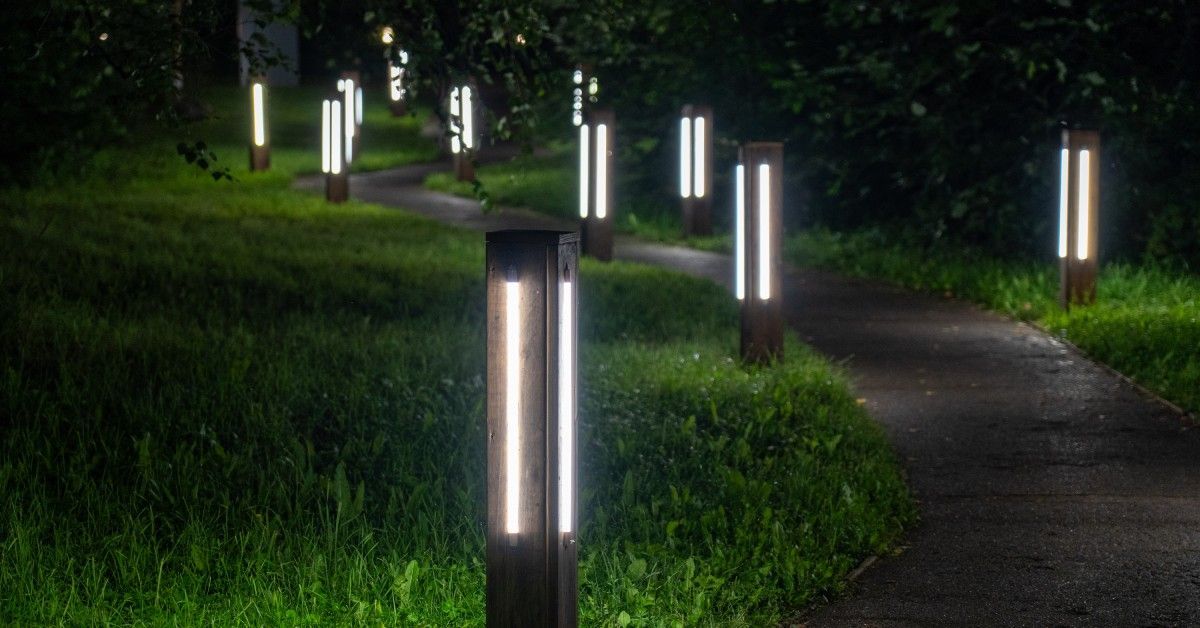 Square safety-lit bollards line a concrete walkway bordered by green grass and scattered trees through the park.