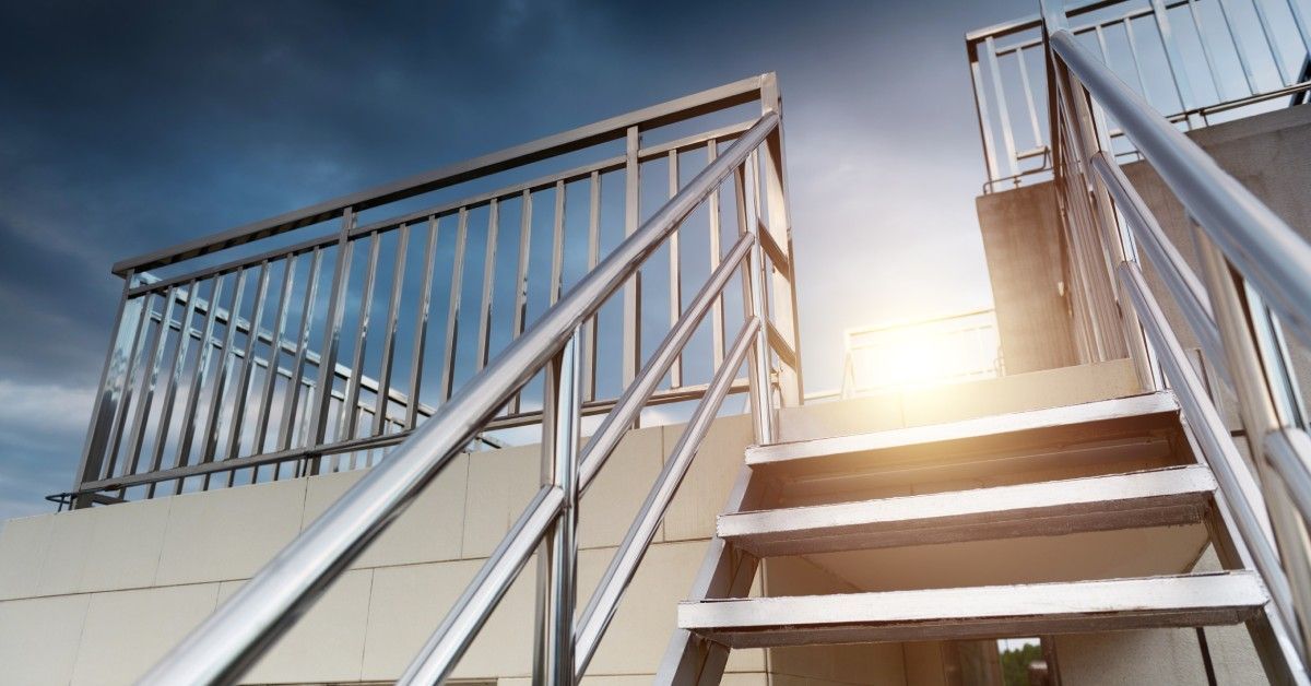 A stainless steel handrail lines a white building with connected stairs, with a warm sunset glowing behind the structure.