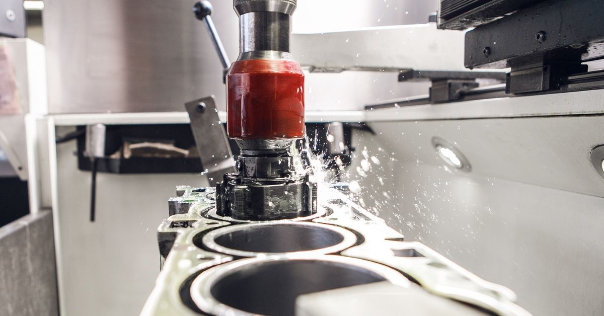A precision milling machine drills into a metal engine block, spraying coolant and fine metallic sha