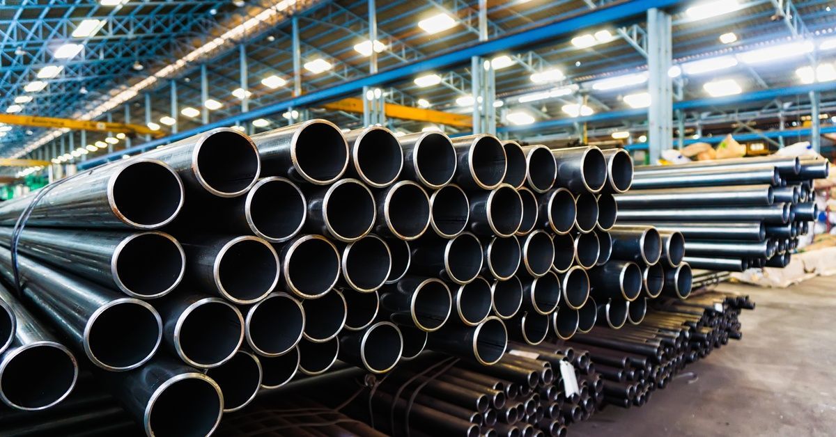 Stacked steel pipes arranged in rows inside large industrial warehouse with overhead lighting and st