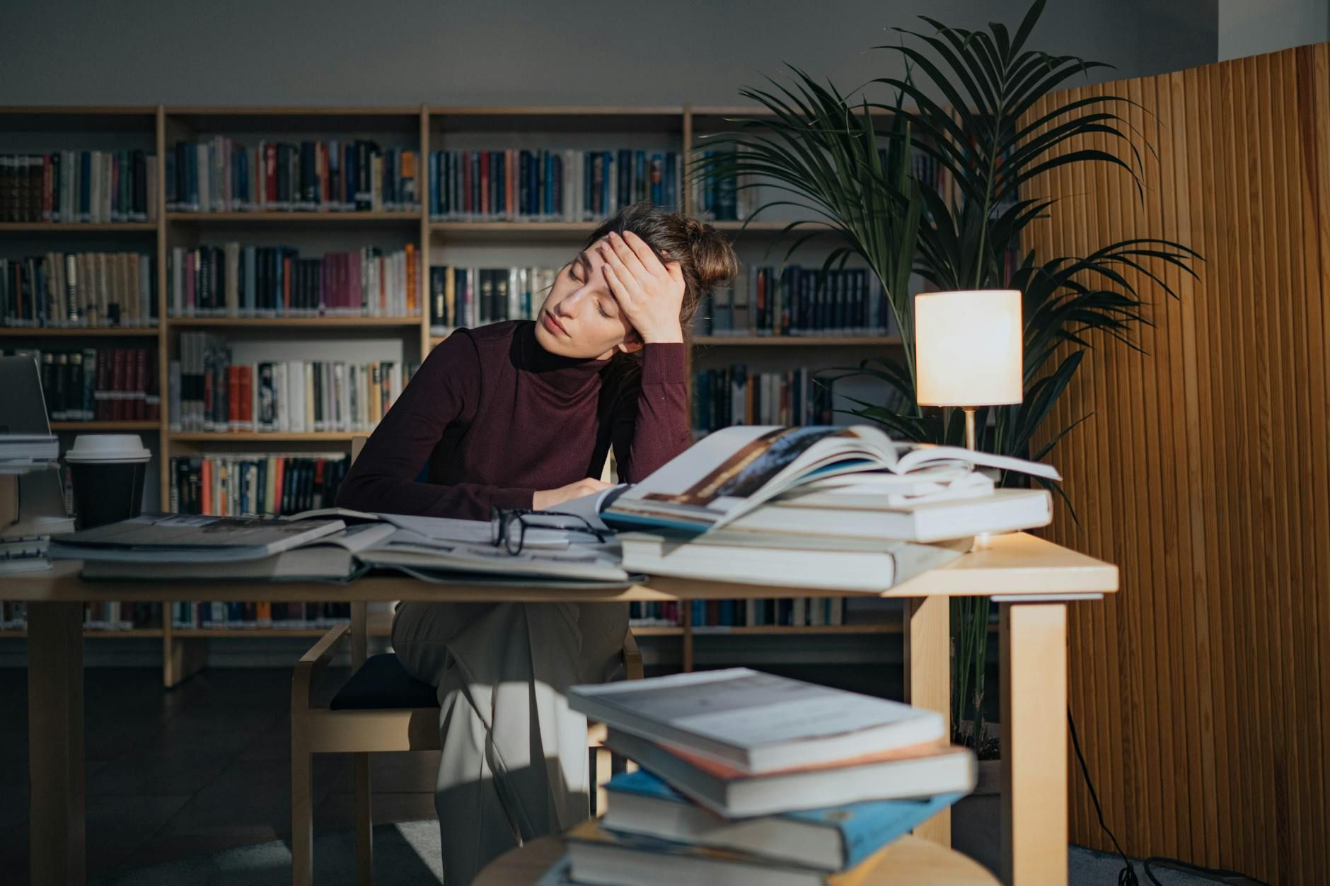 BCBA student in library looking tired while studying, surrounded by open books and a lamp in MD.