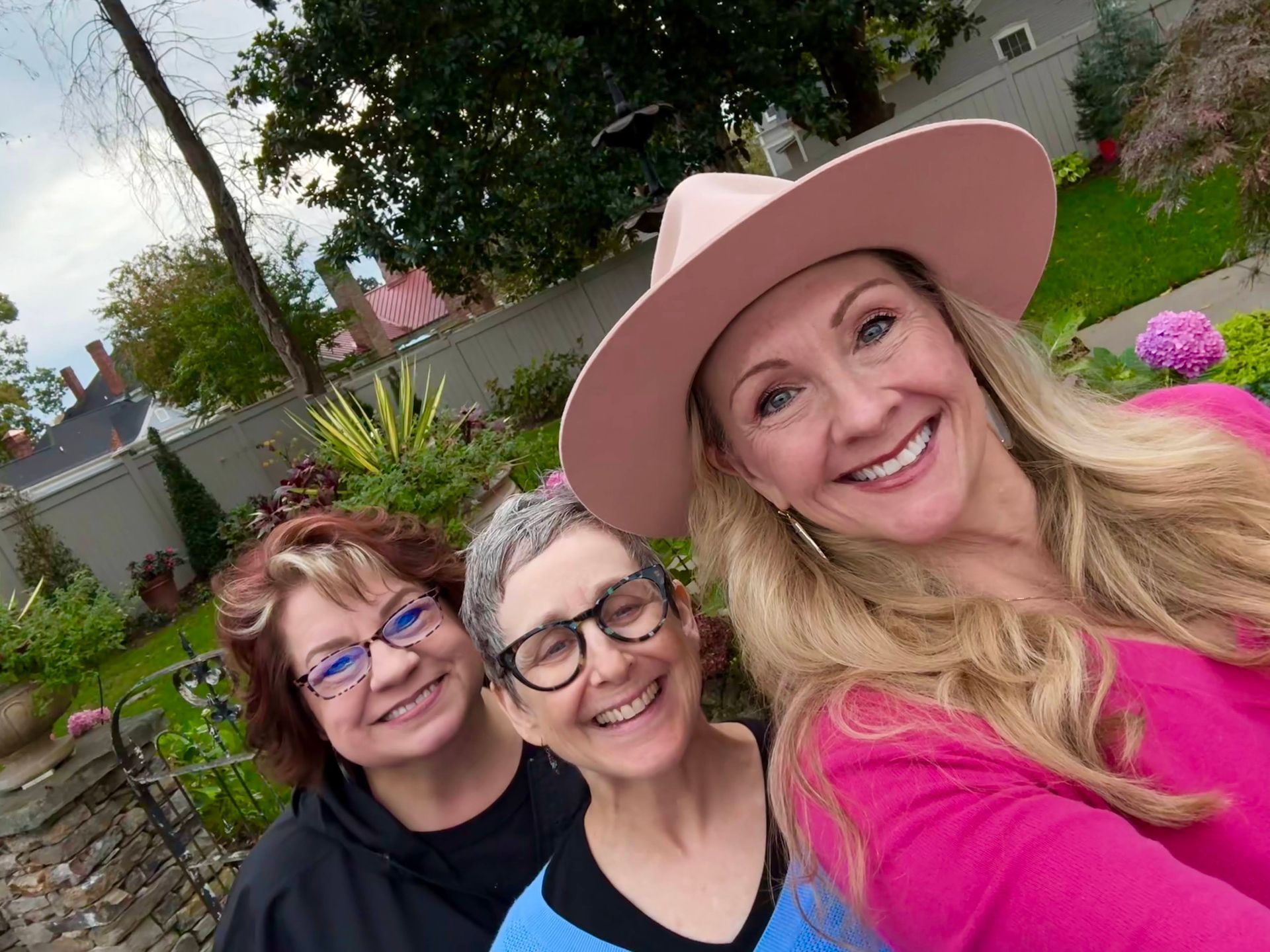 Three women are posing for a selfie in a garden . one of the women is wearing a pink hat.