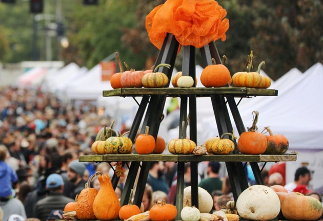 Pumpkin display at a fall festival with crowds, pumpkins of various colors on tiered shelves.