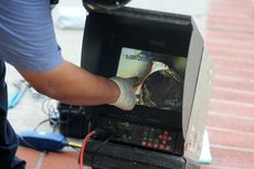 Plumber pointing at a monitor displaying a camera view inside a pipe. Outdoor setting on brick.