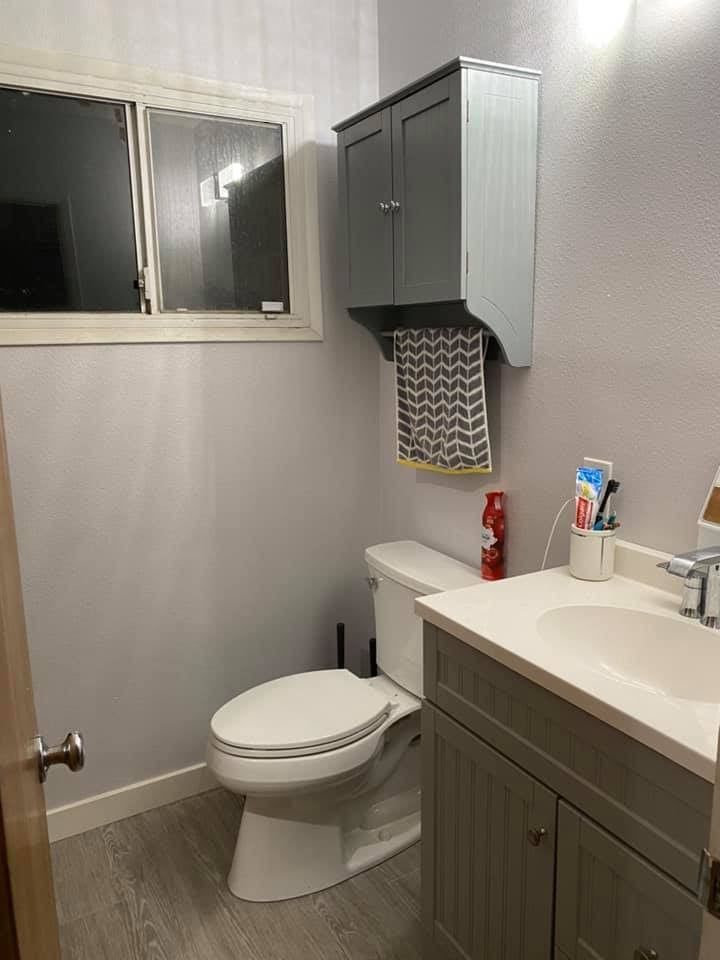 A bathroom with a toilet , sink , cabinet and window.