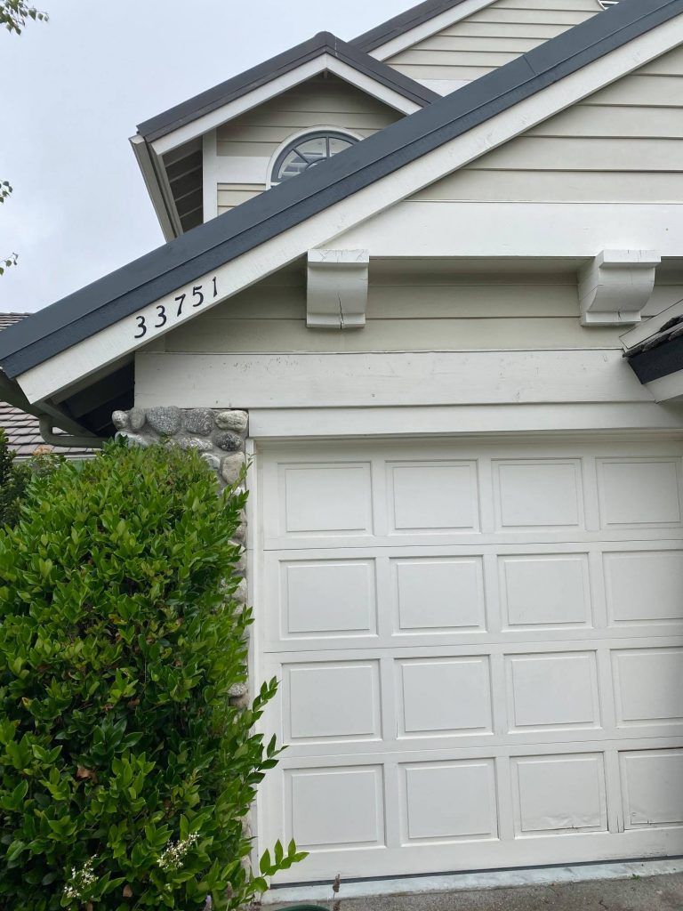 A white garage door is in front of a house with a black roof.