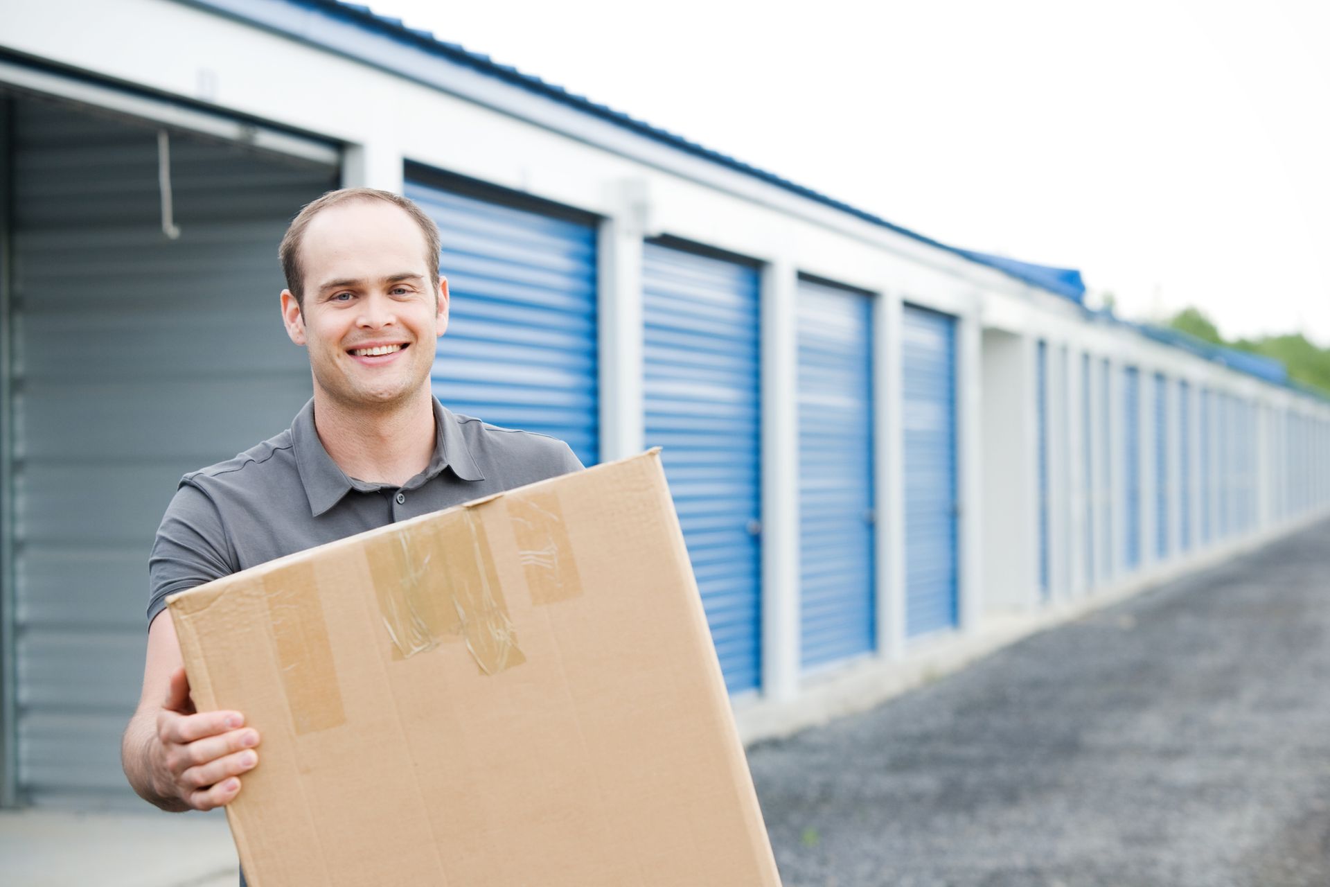 Man holding a large cardboard box smiles in front of blue storage units.
