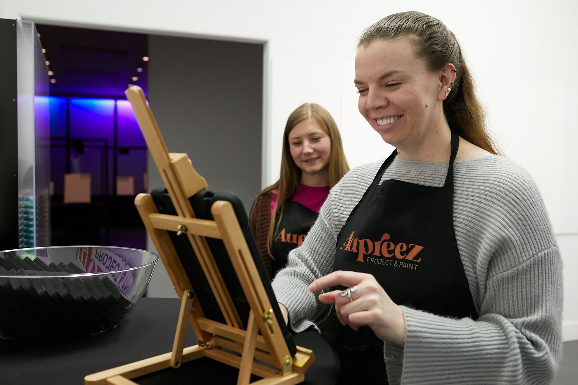 A woman in an apron is standing in front of an easel.