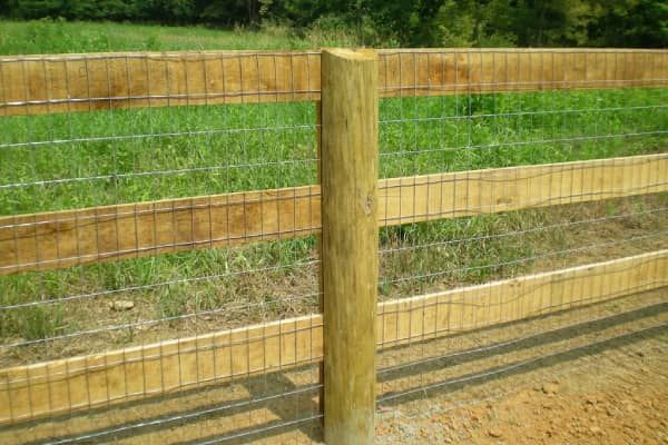 Wooden fence with posts, horizontal boards, and wire mesh in a grassy field.